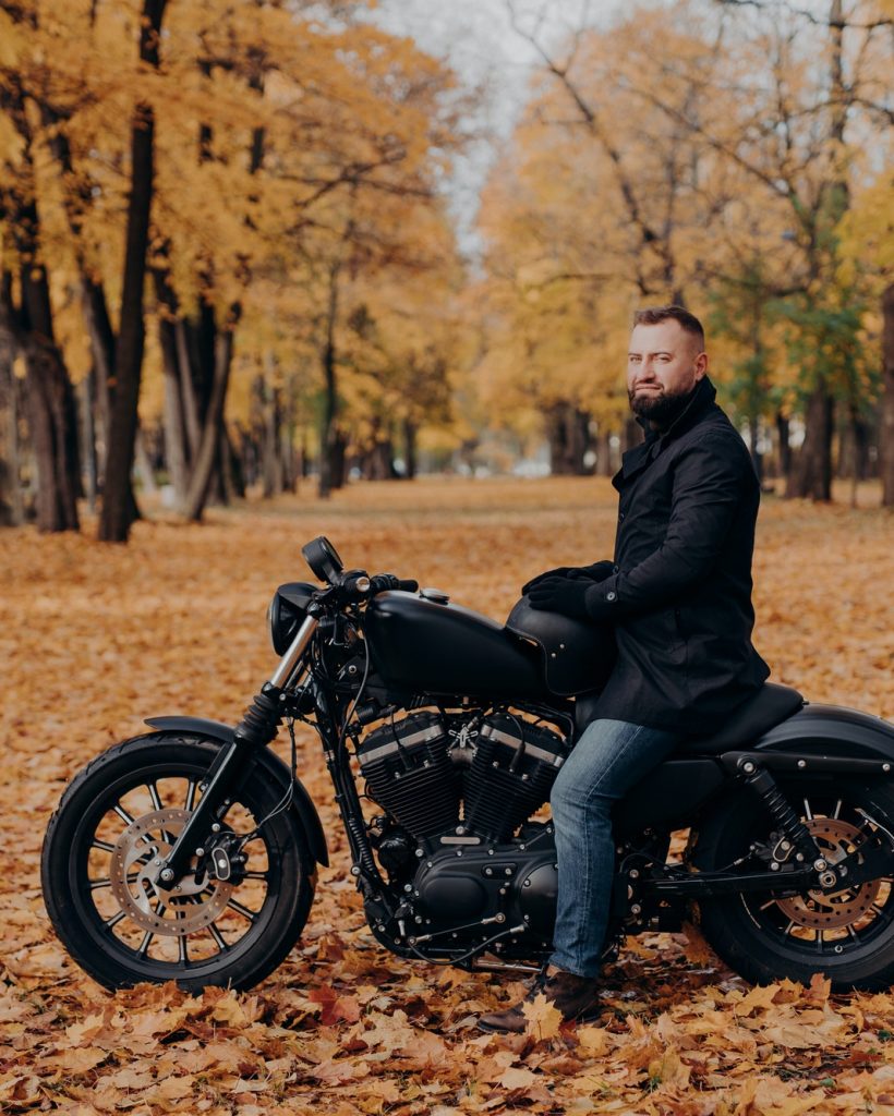 Male biker with thick beard, wears protective gloves, black coat and jeans, poses on motobike.